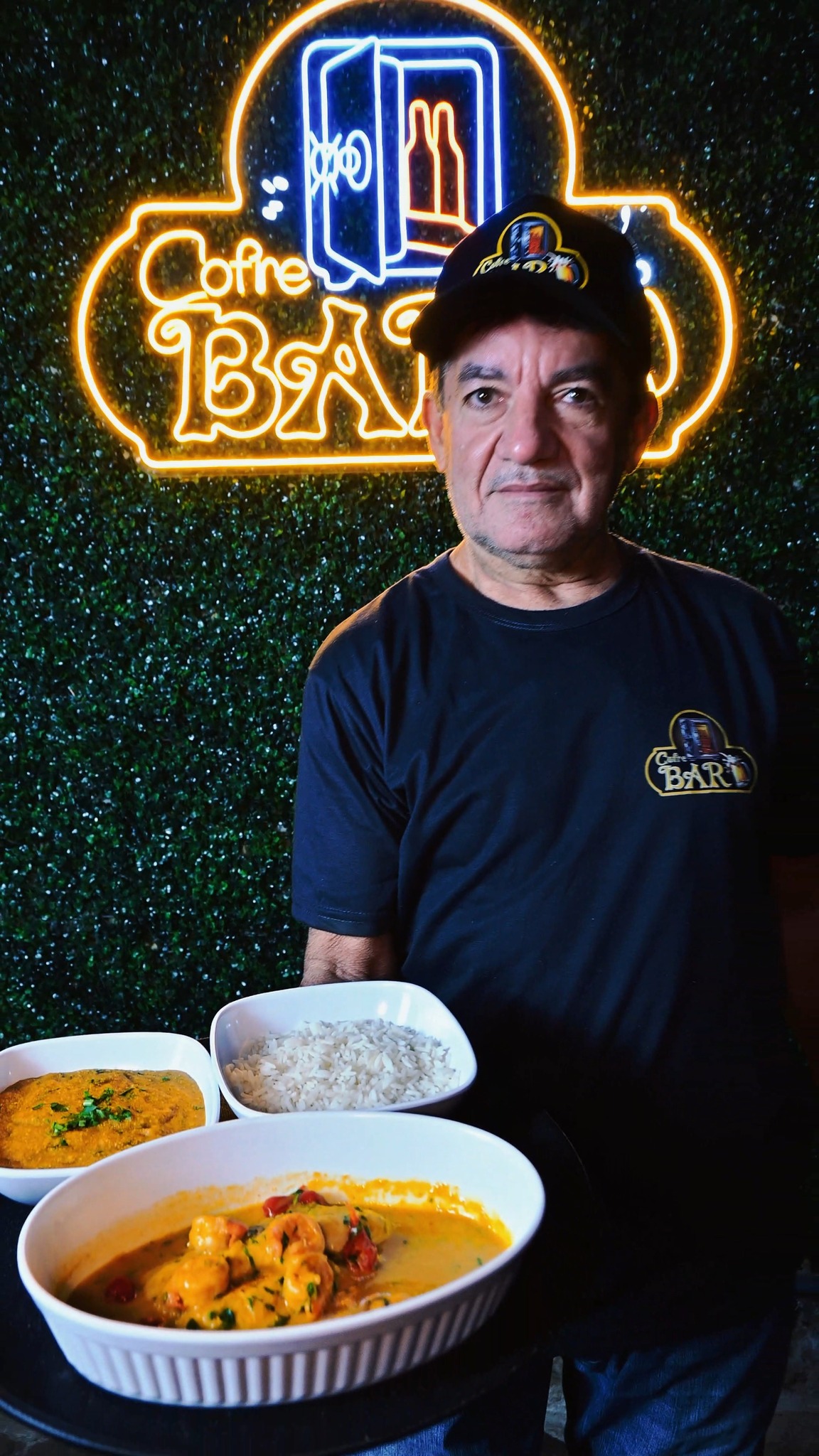 Equipe do Cofre Bar apresentando moqueca de camarão e arroz diante do letreiro de neon